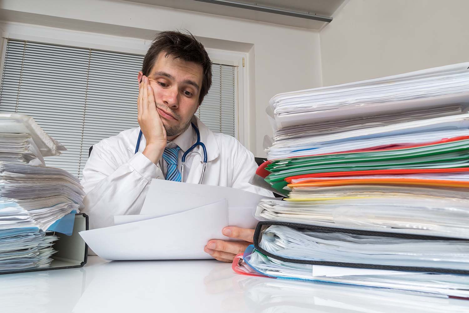 Bureaucracy in medicine concept. Tired overworked doctor is reading medical  report. Many documents on desk. – Breg, Inc.